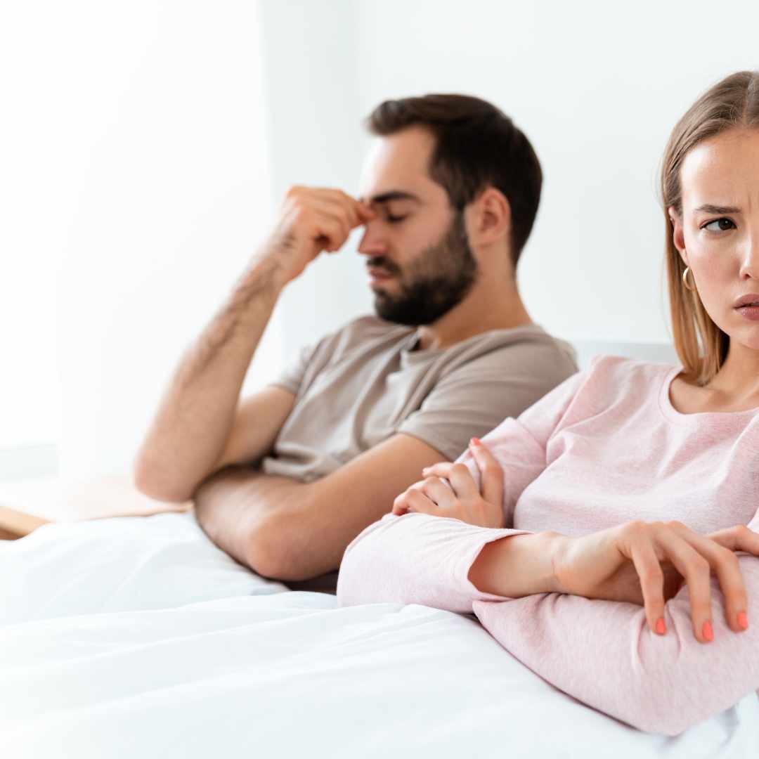 Par ligger adskilt i sengen, kvinde ser frustreret ud, mand holder sig til panden – antyder parproblemer og impotens.