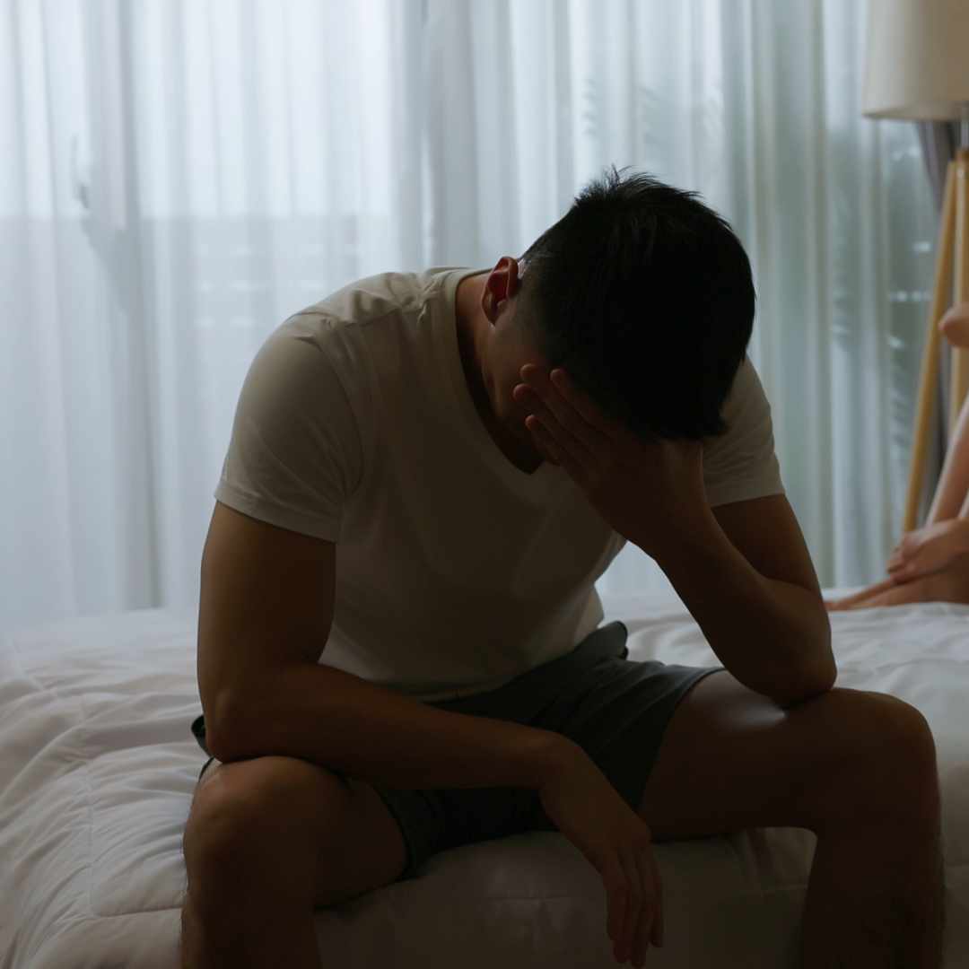 Mand i hvid T-shirt sidder foroverbøjet på sengekant med hånden for ansigtet, symboliserer frustration og bekymring over erektionsproblemer.
