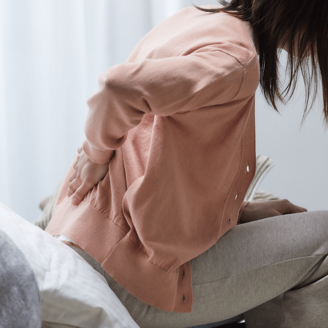 Person der sidder på sengekanten med hånden på lænden og tydelige smerter – et realistisk billede af symptomer ved prolaps i ryggen.