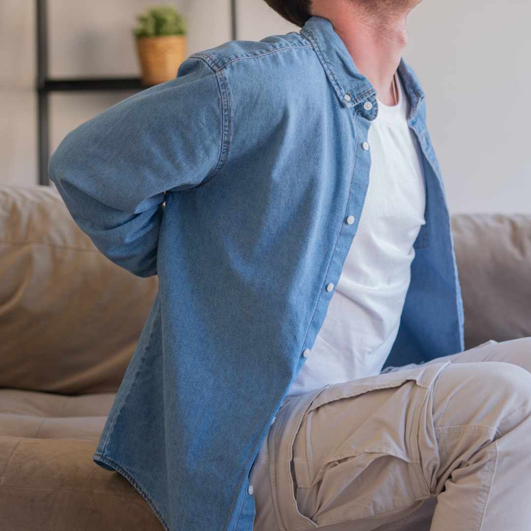 Mand sidder på sofa og holder sig på lænden ved stivhed i kroppen og smerter i ryggen.